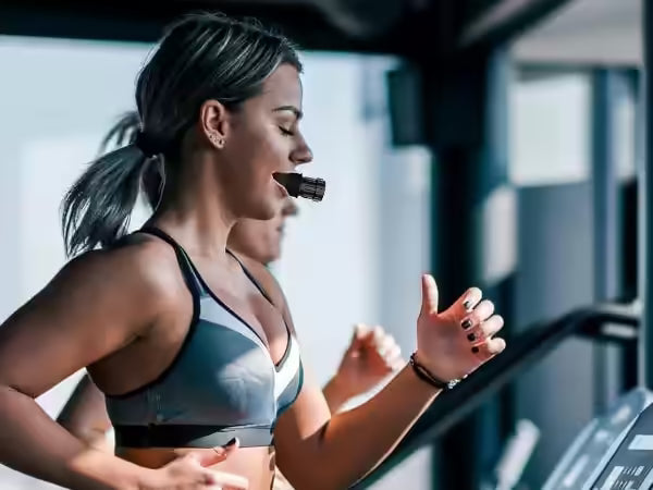 Mujer corriendo en la cinta del gimnasio con el entrenador de respiración en la boca